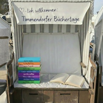 Bild: Ein Strandkorb, über dem steht “Herzlich willkommen: Timmendorfer Büchertage.”. Auf der Sitzfläche liegt rechts ein aufgeschlagenes Buch in Brailleschrift, links ein Haufen verschieden farbiger Bücher von AutorInnen, die bereits bei uns gelesen haben (siehe Rückblick).